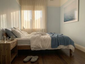 Rustige slaapkamer met wit beddengoed, natuurlijk licht door gordijnen, lavendel op nachtkastje en pantoffels op houten vloer