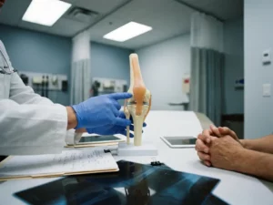 Dokter wijst naar kniegewricht anatomisch model tijdens medisch consult met patiënt, röntgenfoto's op tafel
